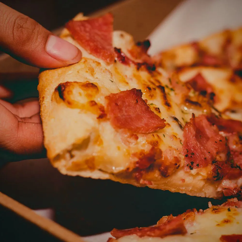 En hånd tar et pizzastykke med skinke og smeltet ost