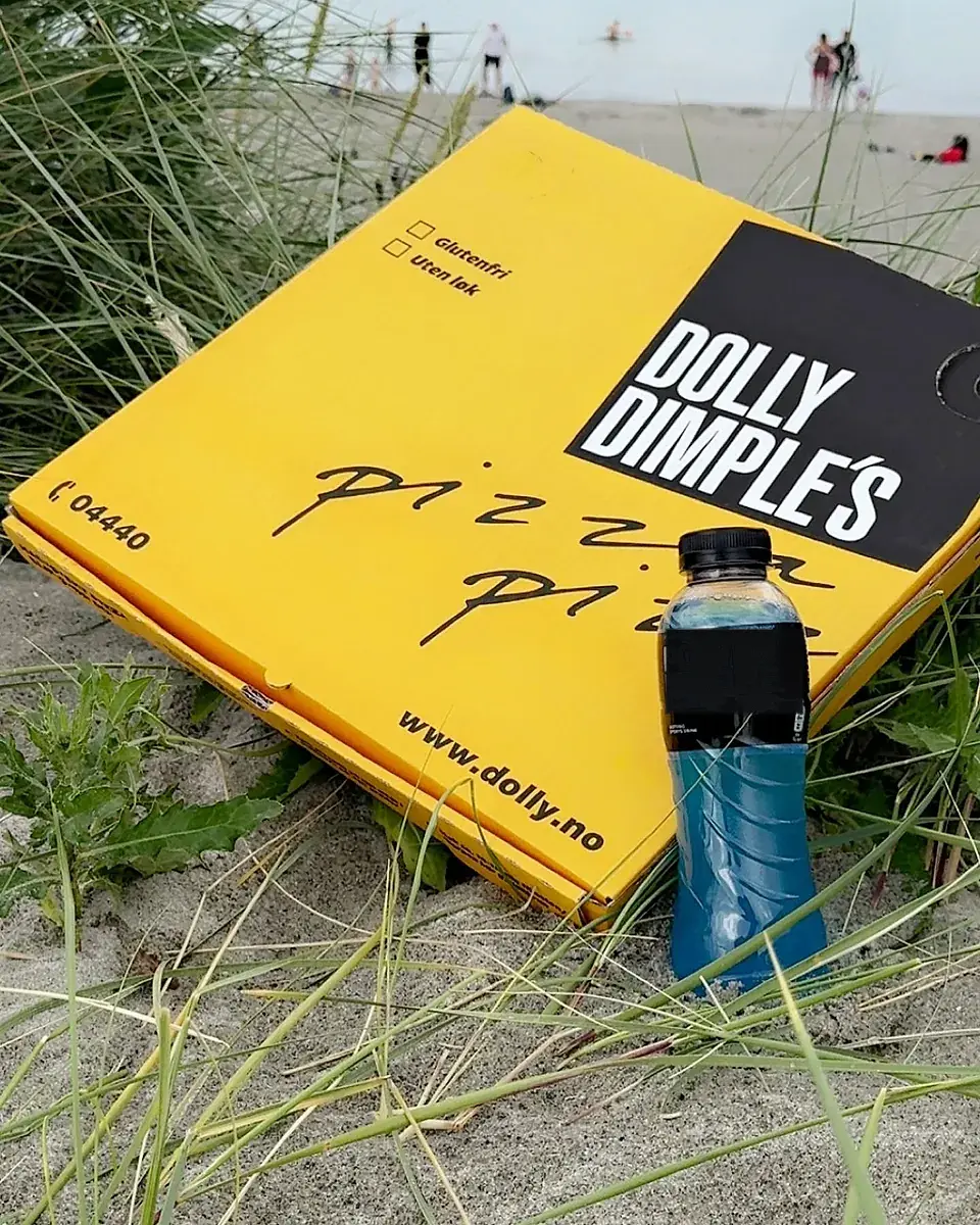 En gul Dolly Dimple’s-pizzaeske i sanddynene på en strand, med en flaske sportsdrikk