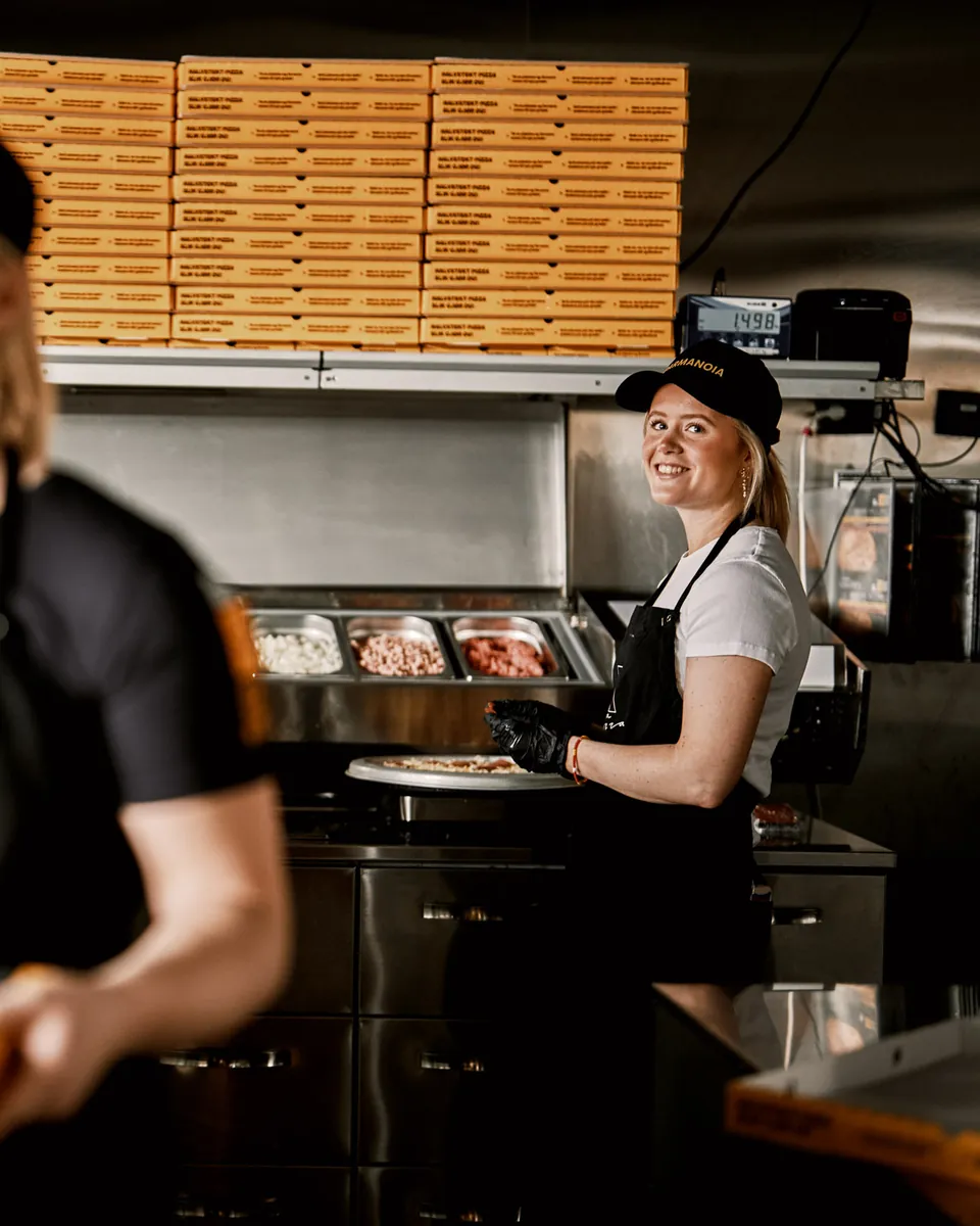 En smilende ansatt på et Dolly-kjøkken, med stabler av pizzaesker bak